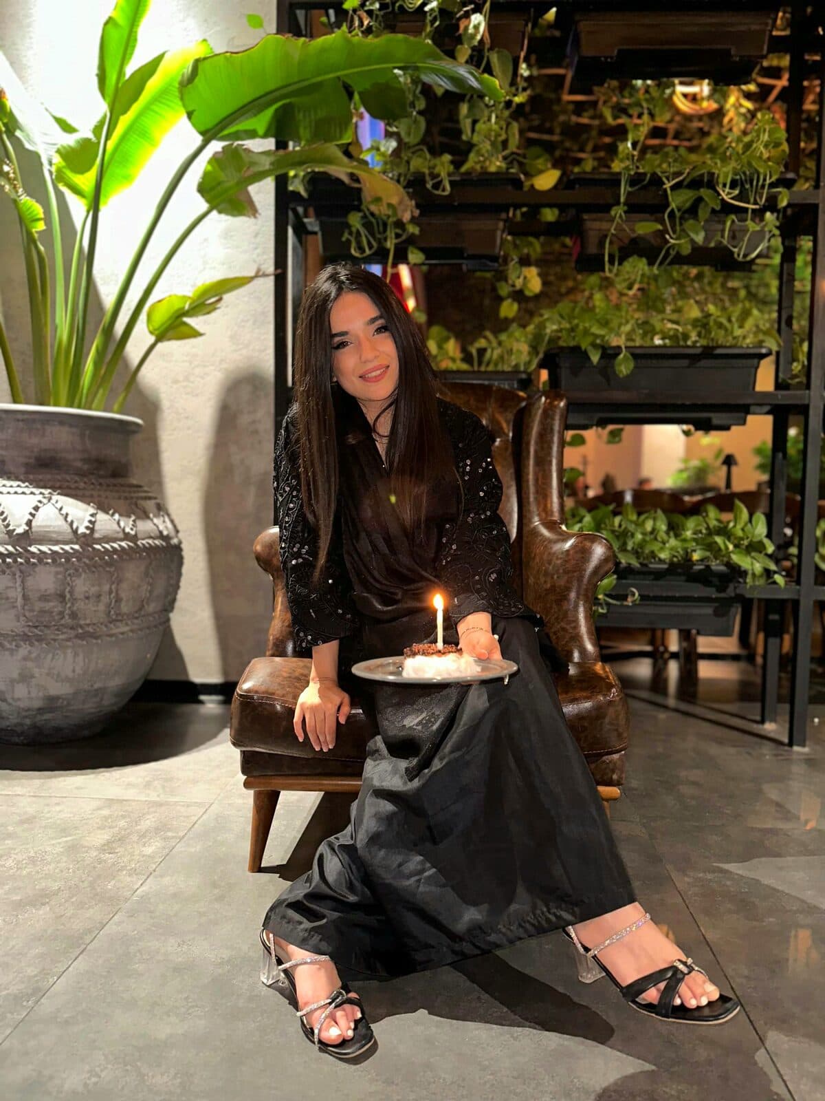 Zahra seated in a leather armchair among plants, holding a small cake with a lit candle