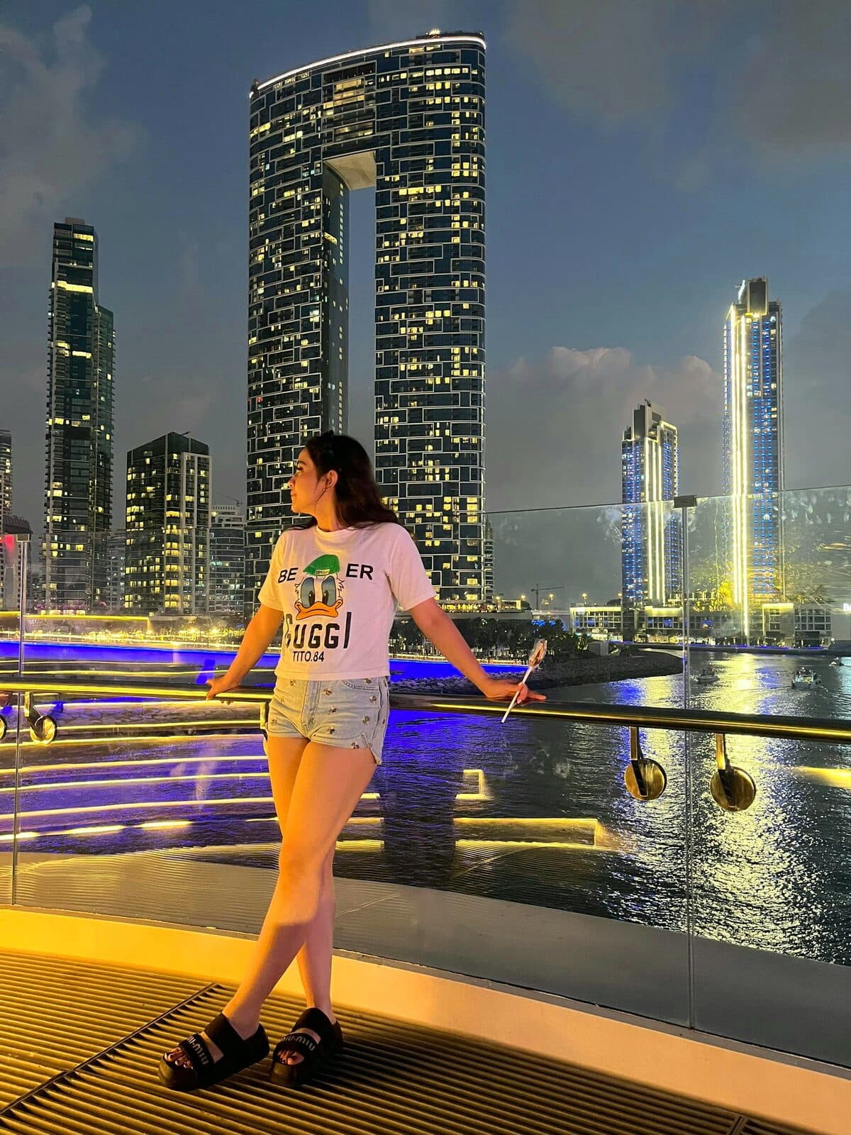 Zahra standing at a waterfront railing with an illuminated skyline behind her